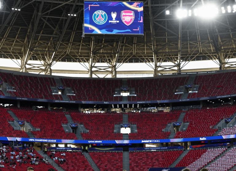 Stadium interior with red seating and a large overhead screen showing PSG and Arsenal logos for a UEFA Champions League final event.