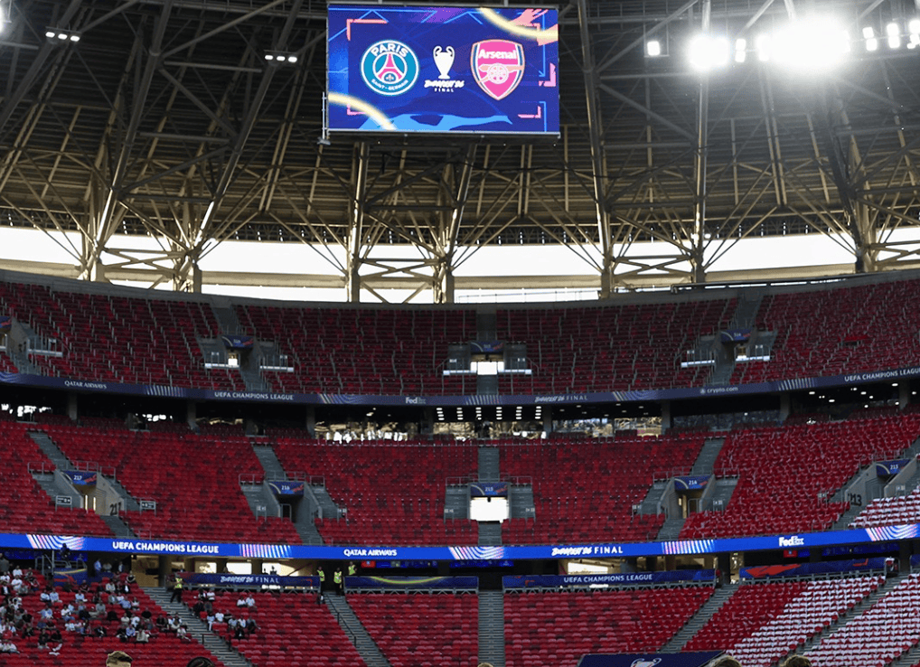 Stadium interior with red seating and a large overhead screen showing PSG and Arsenal logos for a UEFA Champions League final event.