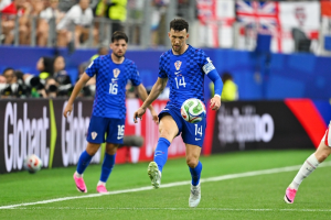 Croatian soccer player #14 controls the ball during a match on a green field, with teammate #16 nearby and fans in the stands behind them.