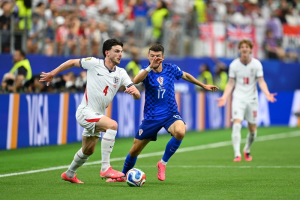 England defender (number 4) in white jersey challenges Croatian midfielder (number 17) in blue for the ball on a soccer pitch during a match with spectators in the background, action-packed scene.