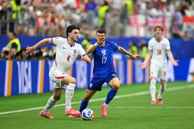 England defender (number 4) in white jersey challenges Croatian midfielder (number 17) in blue for the ball on a soccer pitch during a match with spectators in the background, action-packed scene.