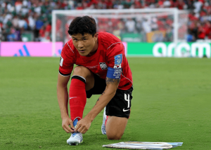 Soccer player in a red jersey kneels on the grass, tying his white cleat before a match with the goal in the background.