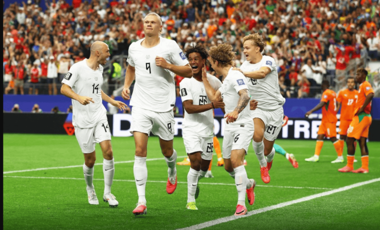 Soccer players in white uniforms celebrate a goal on a green field with a cheering crowd behind them.