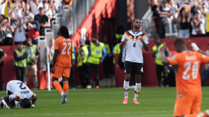 Germany player #4 in white kit stands on the field with fists raised as orange-clad opponents and a fallen teammate are nearby.