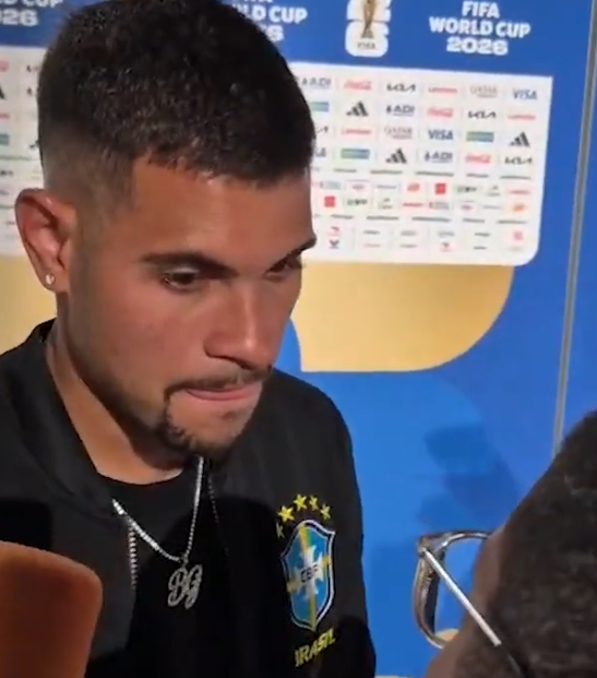 Close-up of a Brazilian footballer with a trimmed beard and earring, wearing a black jacket with the Brazil crest, at a press area with World Cup 2026 branding in the background.