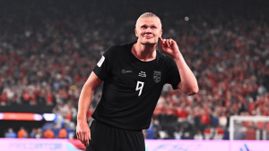 Soccer player in a black jersey with number 9 celebrates on the field in a packed stadium, cupping his ear.