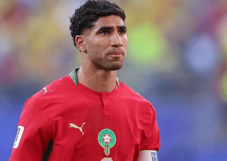 Soccer player in a red Morocco national team jersey with a green crest, looking pensively into the distance with a blurred stadium behind him.
