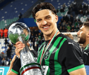Smiling football player in a green and black jersey holding a large trophy with a globe on top, stadium stands in the background and a silver medal around his neck.