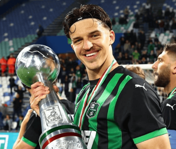 Smiling football player in a green and black jersey holding a large trophy with a globe on top, stadium stands in the background and a silver medal around his neck.