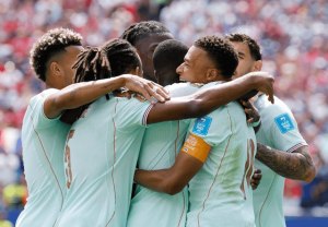 Soccer players in light-blue jerseys form a group hug on the field, captain's orange armband visibleığın.