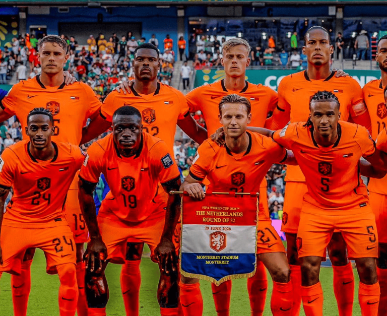 Nederland national football team players in orange kits pose for a group photo on the pitch, holding a banner that reads 'The Netherlands' and shows the Netherlands crest above a date and stadium name.