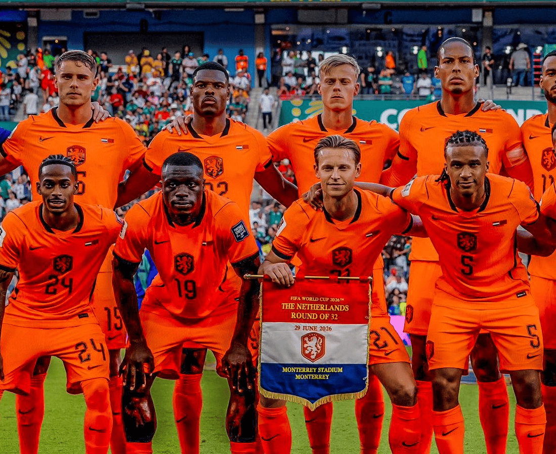 Nederland national football team players in orange kits pose for a group photo on the pitch, holding a banner that reads 'The Netherlands' and shows the Netherlands crest above a date and stadium name.