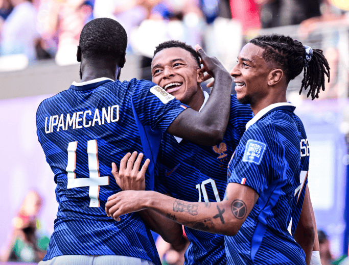 Football players in blue jerseys celebrate a goal, embracing and smiling in a crowd-filled stadium.