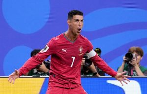 Cristiano Ronaldo in a red Portugal jersey (#7) celebrates with arms wide open on the pitch, photographers in the background in blue stadium scenery.