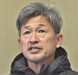 Asian man with gray hair and light stubble wearing a black puffer jacket, speaking in a neutral indoor setting?