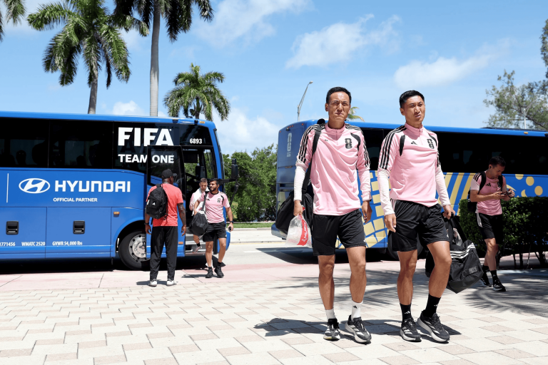 Soccer players in pink training uniforms exit a blue FIFA team bus on a sunny day with palm trees in the background.