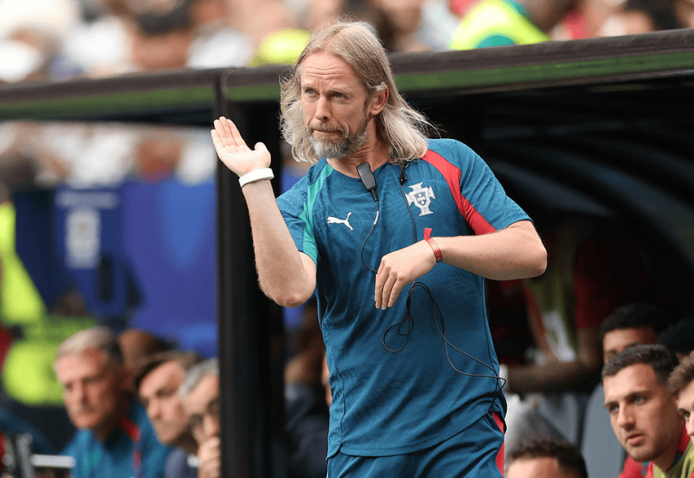 Soccer coach with long gray hair and beard in a blue Portugal training kit, waving from the dugout during a match.