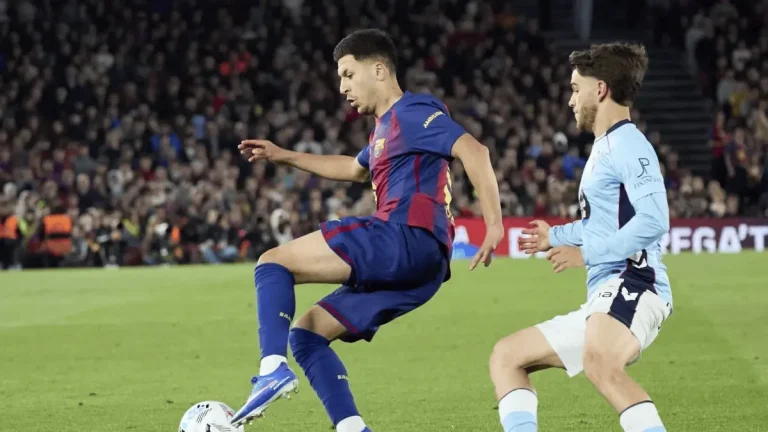 Barcelona player in blue and maroon dribbles the ball as an opposing player in light blue defends on a crowded stadium field.