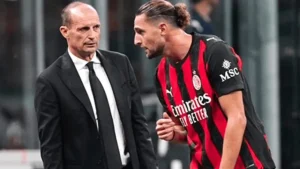 Two men converse on a football pitch: a suited man facing an AC Milan player in red-and-black stripes with long hair tied back.