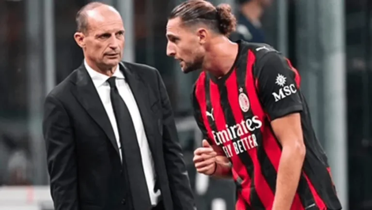 Two men converse on a football pitch: a suited man facing an AC Milan player in red-and-black stripes with long hair tied back.