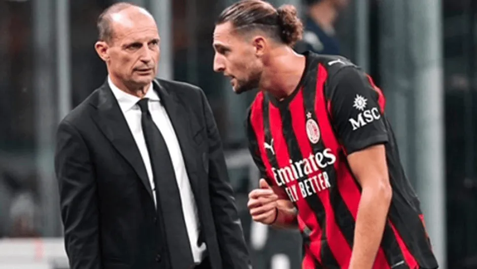 Two men converse on a football pitch: a suited man facing an AC Milan player in red-and-black stripes with long hair tied back.