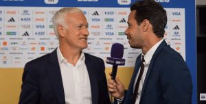 Two men in suits interview each other; reporter holds a purple BeIN Sports microphone in front of a sponsor backdrop.