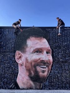 Two people sit on the wall edge of a giant mural portrait of Lionel Messi against a clear blue sky.