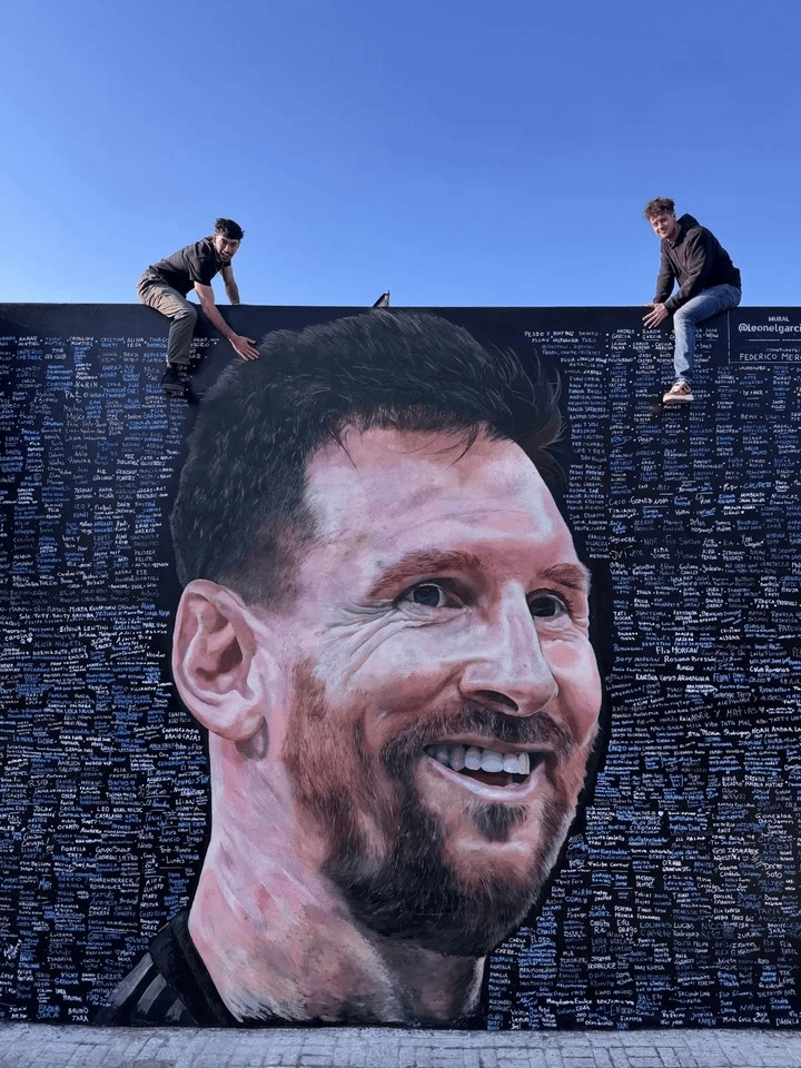 Two people sit on the wall edge of a giant mural portrait of Lionel Messi against a clear blue sky.