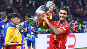 Footballer in red lifts a large silver trophy in celebration, ribbons trailing, as a teammate wrapped in a blue-yellow flag looks on.