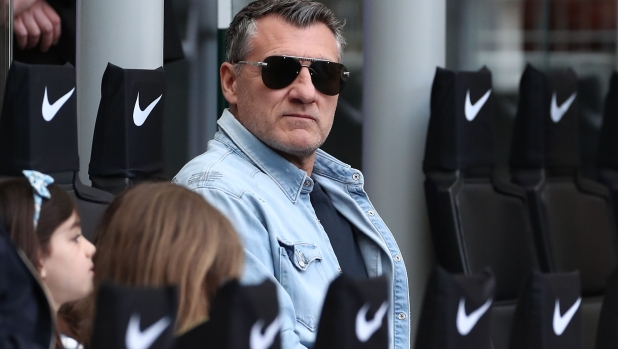 Man wearing a light denim jacket and dark sunglasses sits among stadium seats, with Nike-branded chair backs in the background.