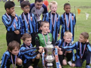 Youth soccer team posing with a large silver trophy and medals on a grassy field after a match.