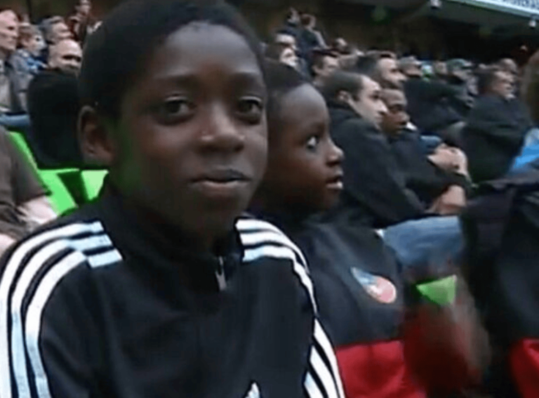 Boy wearing a black Adidas jacket with white stripes sits in a crowded stadium audience, looking toward the camera.