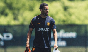 Male soccer player in a dark blue training kit walking on a grassy field, ING sponsor logo visible on chest, holding gear bags nearby.
