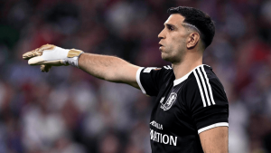 Soccer goalkeeper in a black kit with white stripes points to the left while wearing white gloves.