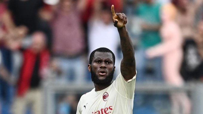 Footballer in a cream AC Milan kit points upward with his right hand in celebration, blurred crowd in the background.