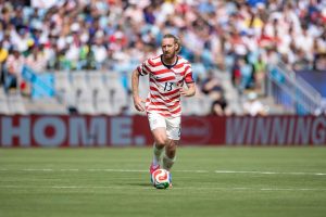 Soccer player in a red-and-white striped USA kit (number 13) dribbles a ball on a green field with a blurred crowd in the background.