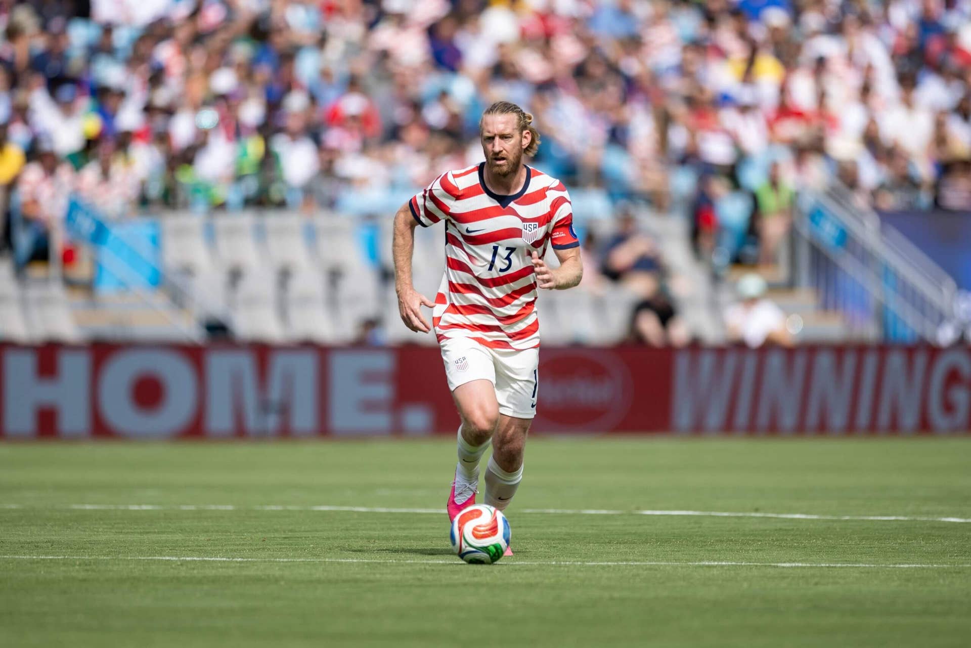 Soccer player in a red-and-white striped USA kit (number 13) dribbles a ball on a green field with a blurred crowd in the background.