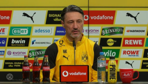 Professional football coach in Borussia Dortmund gear speaks at a press conference behind a sponsor-filled backdrop and microphones on the table.