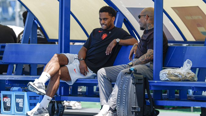 Two men sit on a blue sports bench in a dugout, chatting casually amid bags and gear nearby.