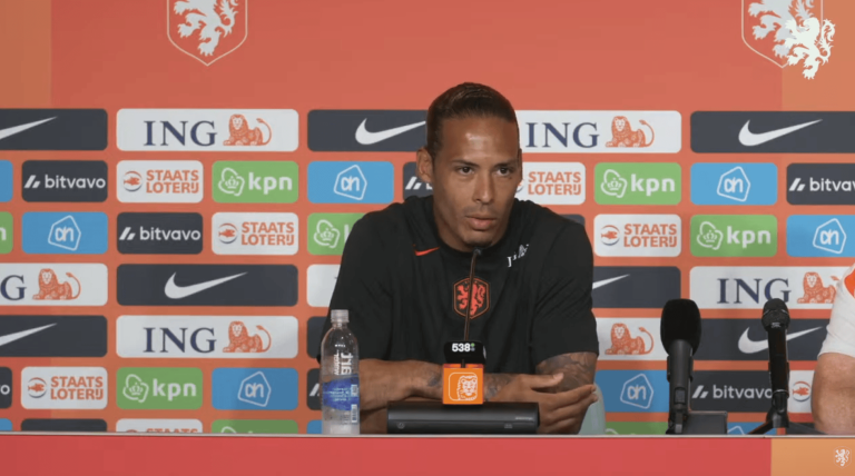 Male football coach at a press conference, seated behind a table with sponsor logos and Dutch crest backdrop.