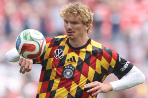 Soccer player in a German national team jersey with a black‑red‑gold diamond pattern juggling a ball in a stadium crowd behind him.