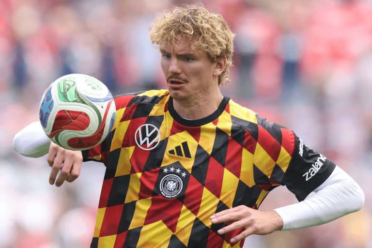 Soccer player in a German national team jersey with a black‑red‑gold diamond pattern juggling a ball in a stadium crowd behind him.