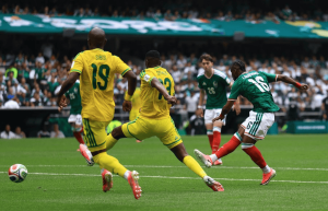 Soccer players in yellow and green clash for the ball on a green pitch with a crowded stadium in the background.