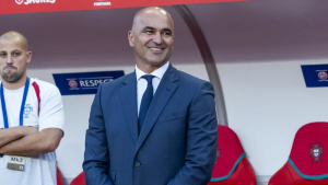 Smiling man in a blue suit stands on a sports bench with red seats behind him, a coach or official at a match.
