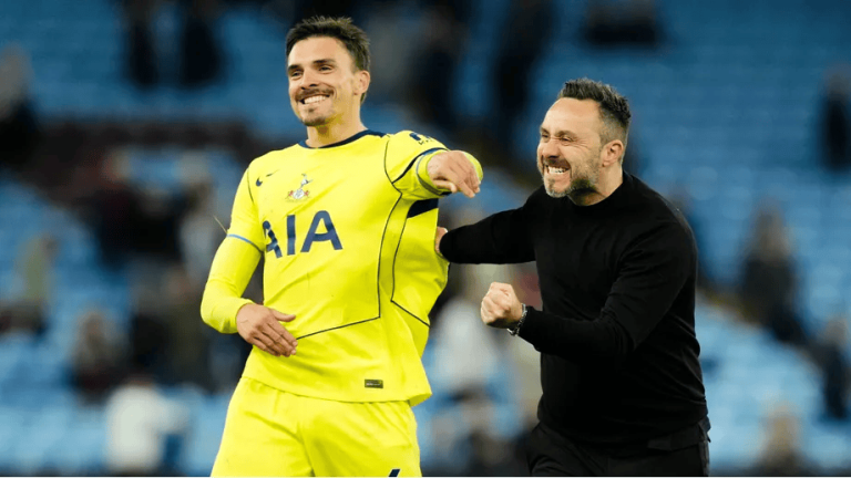 Two men celebrate on a football field, one in a bright yellow kit and the other in black, smiling and linking arms together.