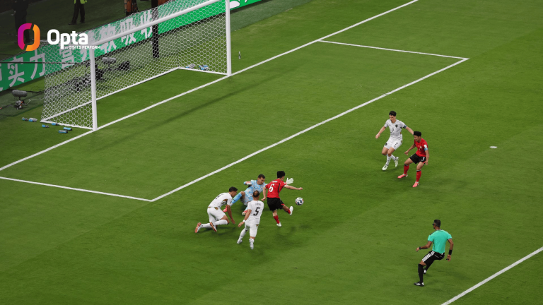 Soccer players in white and red clash near the goal as a shot is taken with the goalie on the ground near the net.
