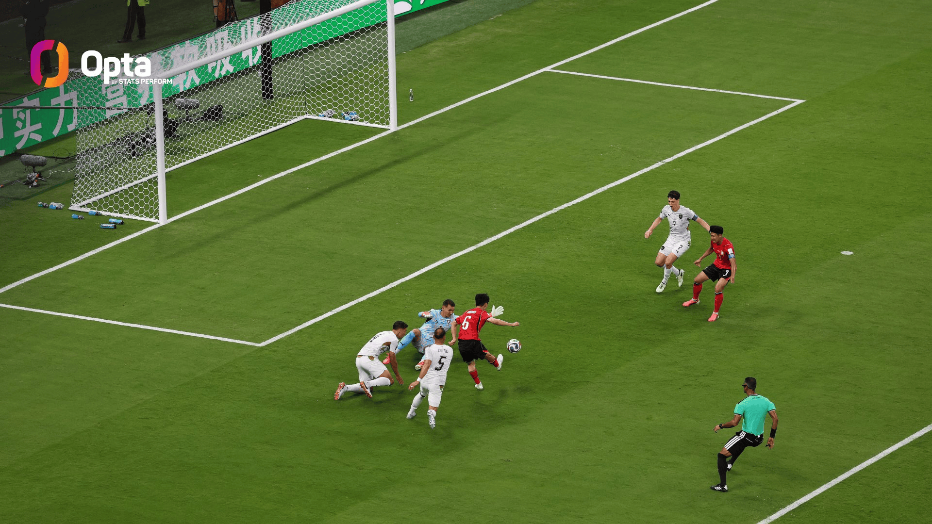 Soccer players in white and red clash near the goal as a shot is taken with the goalie on the ground near the net.