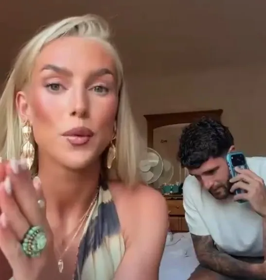 Blonde woman wearing large gold earrings and rings speaks to the camera; a man in a white shirt sits on the bed talking on a blue smartphone in the background.
