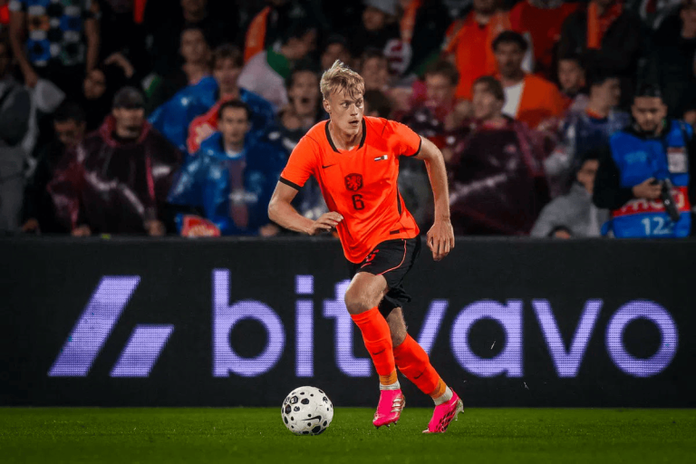 Soccer player in an orange kit with number 6 dribbling the ball on a green field as fans watch from the stands.