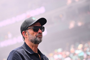 Bearded man in a dark cap and sunglasses at an outdoor event, with a blurred stadium crowd in the background.